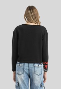 Schwarzes Langarmshirt mit weiter Ausschnittöffnung und rotem Text auf dem Ärmel, kombiniert mit hellblauen Cargo-Hosen mit mehreren Taschen.