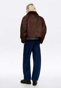 Blondynka stojąca tyłem do kamery, ubraną w brązową skórzaną kurtkę z kożuszkiem, ciemnoniebieskie dżinsy i brązowe buty na białym tle.