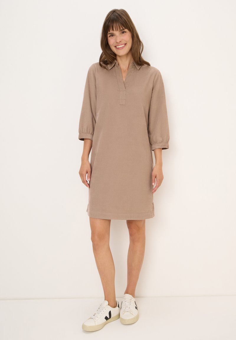 Femme souriante, portant une robe marron ample arrivant au genou avec des manches 3/4 et des baskets blanches, se tenant devant un fond blanc uni.