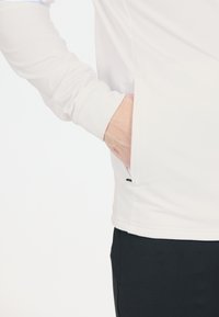 Hvid langærmet top med en glat tekstur, der har en sidelomme og et minimalistisk design. Sæt sammen med mørke bukser, der giver kontrast.