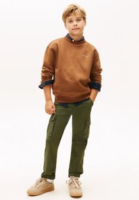 Felpa marrone con polsini a coste, sopra una camicia a quadri blu. Pantaloni cargo verde oliva con tasche, scarpe beige con lacci bianchi.