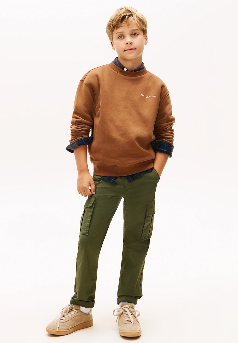 Felpa marrone con polsini a coste, sopra una camicia a quadri blu. Pantaloni cargo verde oliva con tasche, scarpe beige con lacci bianchi.