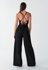Black jumpsuit with wide legs and a back design featuring crisscross straps and a tied accent. Smooth fabric, minimal texture.