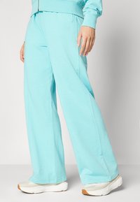 Ανοιχτό μπλε φόρμες από μαλακό μείγμα βαμβακιού, με άνετη εφαρμογή, ζώνη στη μέση και μικρό λογότυπο μπροστά. Συνδυάζεται με λευκά αθλητικά παπούτσια.