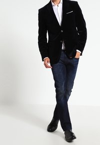 Svart sammetsblazer med slagkrage, knappstängning, i kombination med mörkblå jeans och svarta skor. Vit skjorta synlig under blazern.