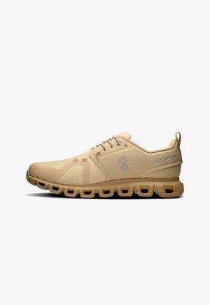 Beige hardloopschoenen met een bovenkant van mesh, een gevoerd kraagje en een vetersluiting. Beschikt over een kenmerkende zool met holle secties voor demping.