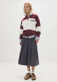 Pullover a righe bordeaux e crema con stampa "NEW YORK CITY", indossato sopra una camicia a righe blu. Gonna plissettata grigia e scarpe marroni con lacci.