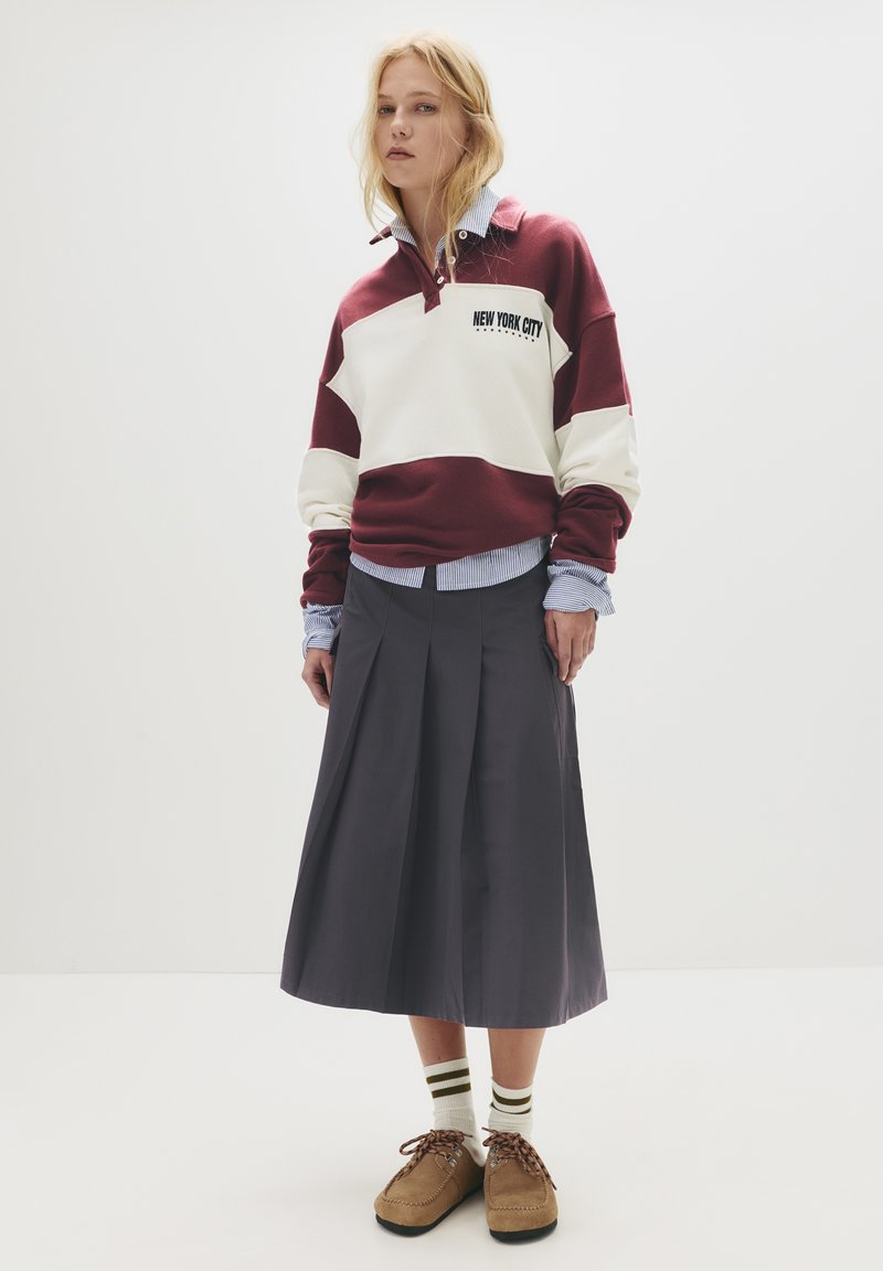 Pullover a righe bordeaux e crema con stampa "NEW YORK CITY", indossato sopra una camicia a righe blu. Gonna plissettata grigia e scarpe marroni con lacci.