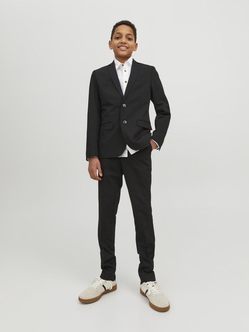 Zwarte suit met een enkelvoudig jasje, wit shirt met knopen, en beige casual sneakers. Slank fit ontwerp, zachte textuur.