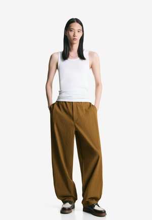 Witte tanktop, bruine wijde plooibroek en twee-kleurige schoenen. De outfit heeft een gestructureerde stof met een relaxte pasvorm en neutrale kleuren.