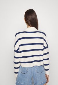 Suéter a rayas en crema y azul marino; textura de punto, diseño corto con mangas anchas, combinado con pantalones cortos de denim con bolsillos traseros decorados.