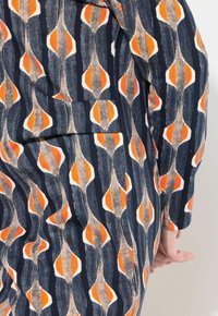 Un tessuto a motivi presenta uno sfondo blu scuro con forme alternate in arancione, bianco e beige. La texture appare morbida e intrecciata.
