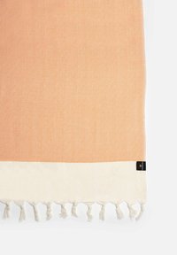Textiel in perzik met een diamantpatroon, crèmekleurige zoom en franjes aan de randen. Etiket zichtbaar in de onderste hoek. Zachte, geweven textuur.
