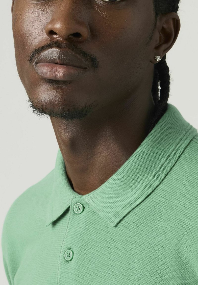 Hombre con cabello trenzado, barba recortada y pendiente de botón, vistiendo una camisa verde claro con cuello y dos botones abrochados.