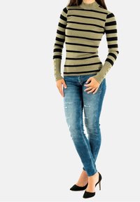 Gestreepte ribben top met lange mouwen in olijfgroen en zwart, gecombineerd met versleten blauwe jeans en zwarte puntige hakken. Gedetailleerde knoopaccenten op de mouwen.