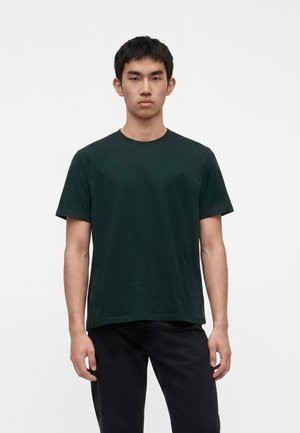 Jeune homme aux cheveux courts et noirs portant un t-shirt vert foncé à manches courtes et un pantalon noir, debout sur un fond blanc uni.