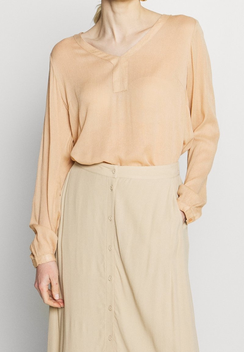 Blouse pêche avec un col en V et des manches longues, fabriquée à partir d'un tissu léger. Assortie d'une jupe beige avec des boutons sur le devant et une texture douce.