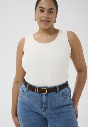 Femme portant un haut blanc sans manches, un jean bleu avec une ceinture marron et des boucles d'oreilles en forme d'anneaux dorés, debout devant un fond clair uni.