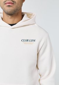 Man bär en off-white hoodie med texten "CLSR LDN Sports & Lifestyle Premium Wear" på övre vänstra bröstet, mot en enfärgad bakgrund.