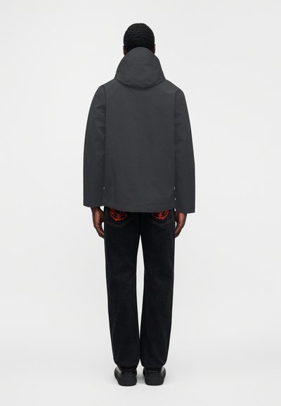 Μαύρο μπουφάν με κουκούλα και κομψό σχεδιασμό, σε συνδυασμό με σκούρο τζιν με κόκκινες κεντητές λεπτομέρειες στις πίσω τσέπες.