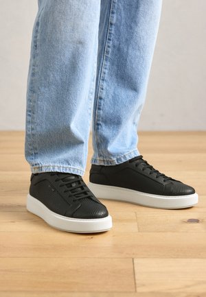 Persoon in lichtblauwe jeans en zwarte sneakers met een gestructureerd patroon en witte zolen, staande op een licht houten vloer.