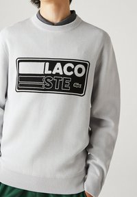 Osoba ubrana w jasnoszary sweter Lacoste z czarno-białą łatką z logo oraz małym zielonym emblematem krokodyla na piersi.