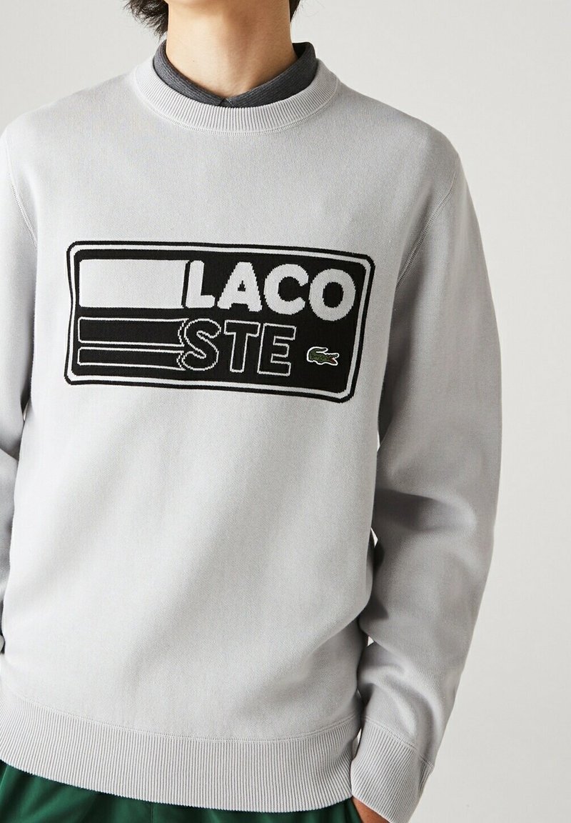 Osoba ubrana w jasnoszary sweter Lacoste z czarno-białą łatką z logo oraz małym zielonym emblematem krokodyla na piersi.