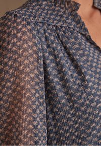 Blouse bleu à motifs floraux avec coutures d'épaules froncées et petits plis près du col sur le haut du torse d'une personne.