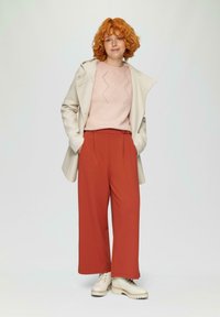 Roze gebreide trui met ruitpatronen, crèmekleurige jas en rode wijde broeken; model draagt beige veterschoenen.
