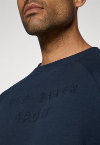 Felpa blu navy con logo goffrato "TED BAKER SPORT." Caratterizzata da un colletto rotondo e maniche raglan. Texture del tessuto liscia.