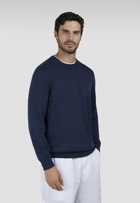 Maglione lavorato a maglia blu navy con scollo a girocollo e polsini a costine, abbinato a pantaloni di lino bianchi. Texture liscia e vestibilità classica.