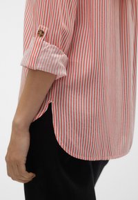 Chemise rayée en coton rouge et blanc avec manches retroussées, détail de boutons, ourlet arrondi et fentes latérales.