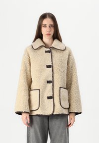 Beige schapenvachtjack met een grote kraag, bruine knoopsluitingen en twee voorzakken met een donkere rand. Relaxte pasvorm en gestructureerd.