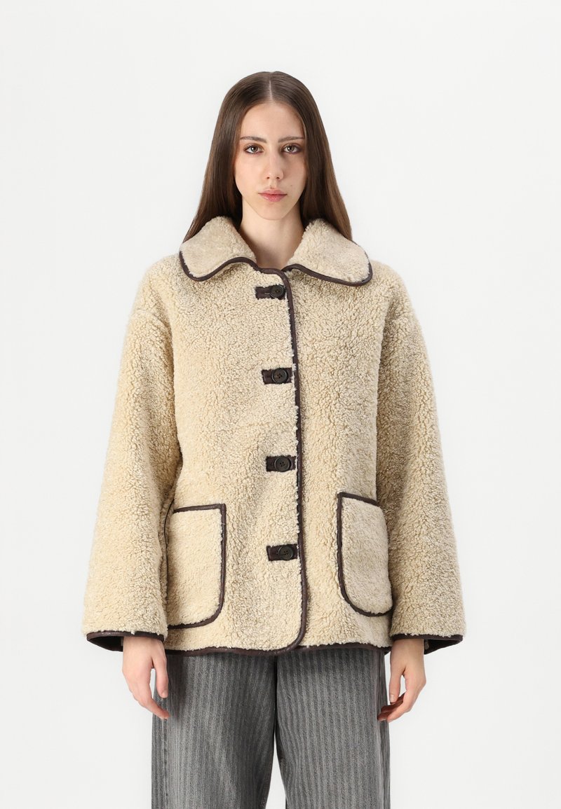 Beige schapenvachtjack met een grote kraag, bruine knoopsluitingen en twee voorzakken met een donkere rand. Relaxte pasvorm en gestructureerd.