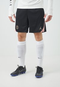 Svarta sportshorts med en rosa detalj, med en guld Paris-logotyp. Vita knähöga strumpor och svarta fotbollsskor med blå sulor.