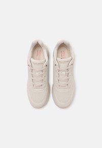 Ett par beige Skechers-sneakers med perforerade tår, snördesign och vadderade innersulor sedda ovanifrån på en vit bakgrund.