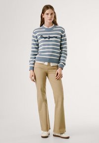 Suéter a rayas azul y blanco con logo, puños acanalados, combinado con pantalones acampanados beige y zapatos blancos. Textura suave, corte casual.