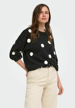 Ung kvinde iført en sort sweater med store hvide polkaprikker og beige høj-taljede bukser, stående med den ene hånd på hoften.