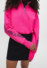 Růžová zipová mikina s ribbeným límcem, na rukávech s potiskem "CALVIN KLEIN JEANS" černou barvou. Pod ní je viditelná černá denimová sukně.