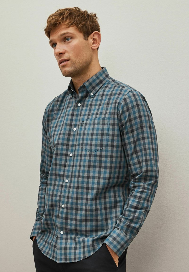 Camisa de manga larga para hombre en un patrón a cuadros azul y gris, con cuello abotonado y un bolsillo en el pecho. Tejido de algodón con una textura suave.