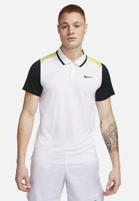 Giovane uomo con capelli corti che indossa una polo bianca Nike con maniche nere, dettagli spalla giallo neon e pantaloni corti bianchi, in piedi contro uno sfondo bianco.