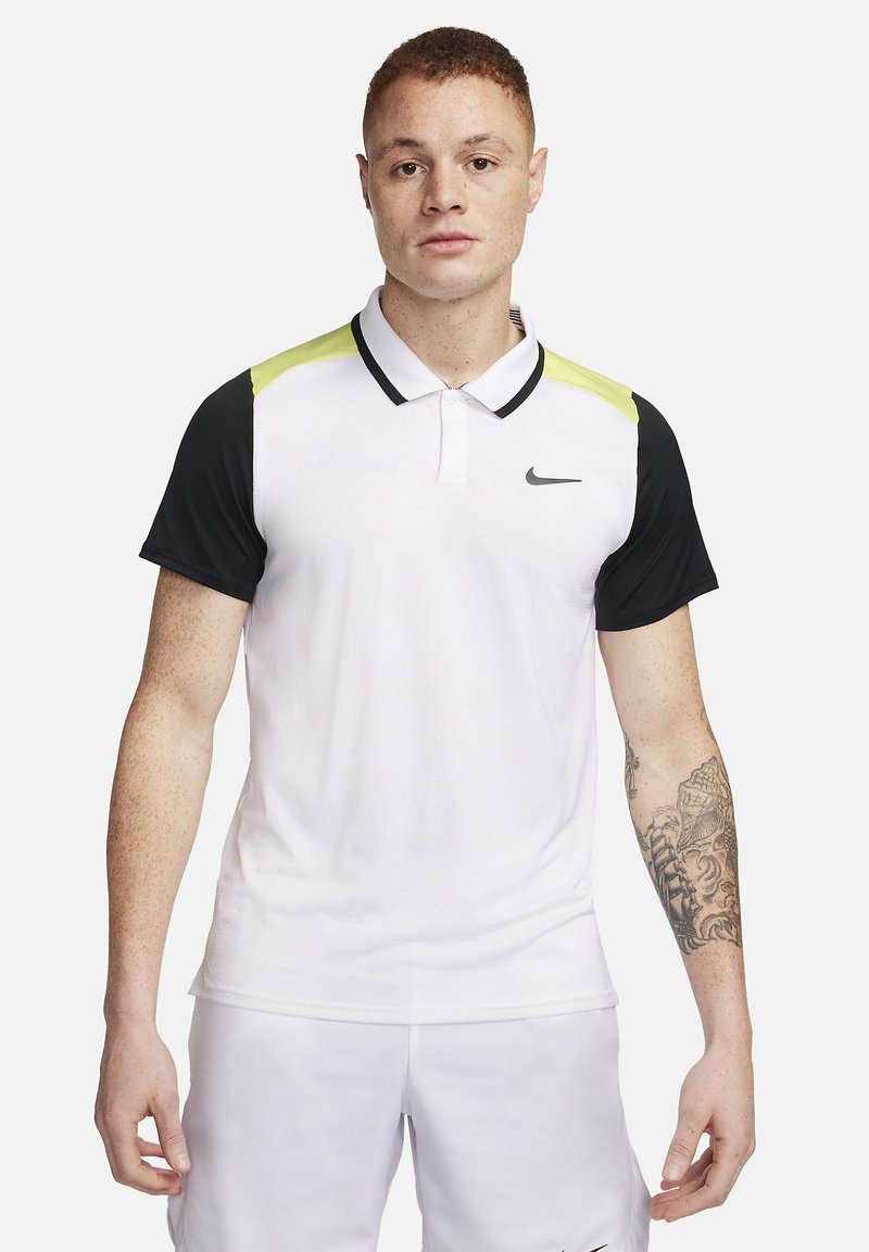 Giovane uomo con capelli corti che indossa una polo bianca Nike con maniche nere, dettagli spalla giallo neon e pantaloni corti bianchi, in piedi contro uno sfondo bianco.