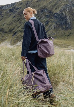 Ung kvinne i mørk genser går gjennom gresset mens hun holder og har på seg matchende lilla vesker i et fjellandskap.