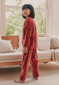 Red velvet tracksuit with puffy sleeves and elastic cuffs. Child standing barefoot on carpet, facing sideways, natural light in background.