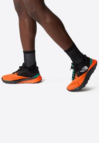 Zapatillas de running con un upper de malla naranja, detalles en negro y un contrafuerte verde, combinadas con calcetines a rayas negros y grises.
