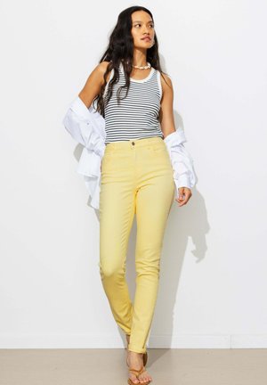 Femme aux longs cheveux foncés portant un débardeur blanc et noir à rayures, un pantalon jaune, un collier de coquillages et tenant une chemise blanche, debout.