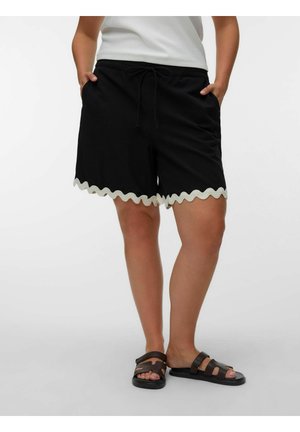 Person iført sorte shorts med hvid bølget kant, hvid top og brune slip-on sandaler, stående med hænderne i lommerne.