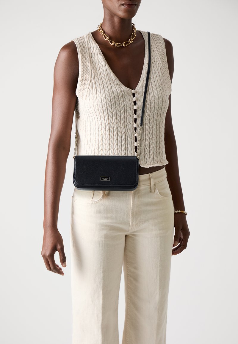 Black leather crossbody bag with a smooth texture, rectangular shape, and gold hardware detail, worn with a cream knit top and light pants.