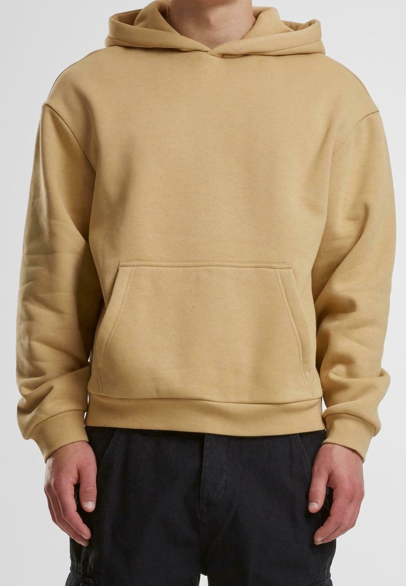 Osoba ve béžové mikině s kapucí, přední kapsou a žebrovanými manžetami, která je spárována s volnými černými kalhotami.