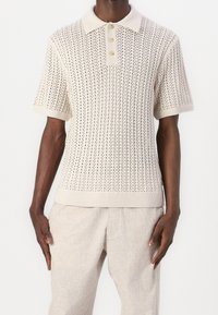 Camisa polo de punto beige con cuello acanalado, mangas cortas y un patrón texturizado con cierre de tres botones.